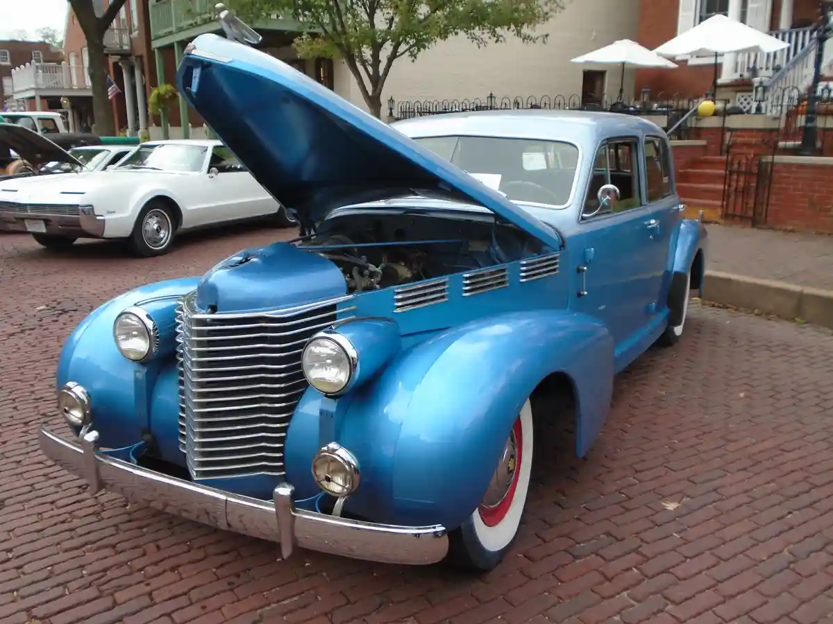 A vintage blue car with its hood open is parked on a brick street, revealing its engine. Another classic car is visible in…