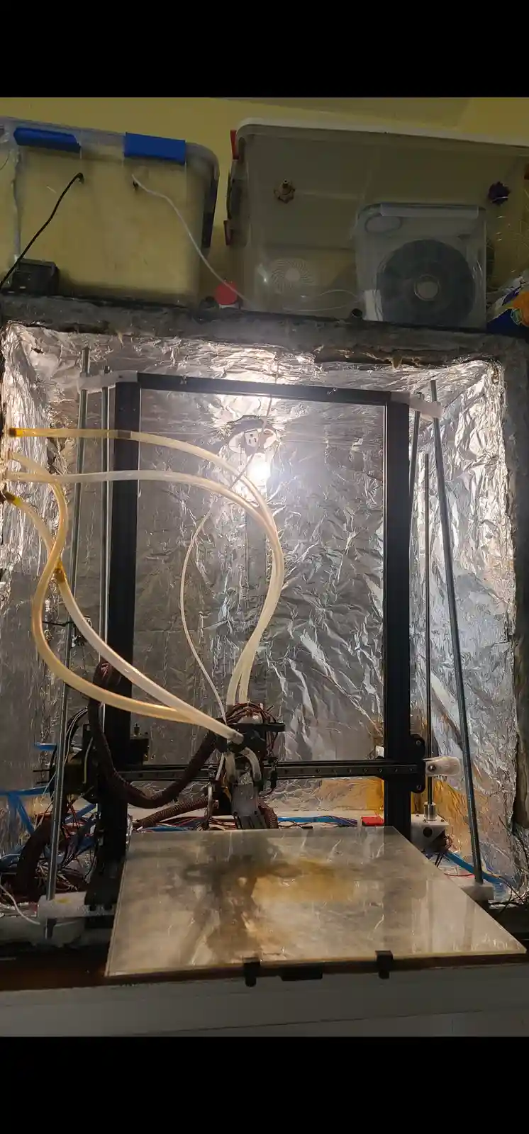 A homemade 3d printer with a transparent build chamber lined with reflective foil, featuring multiple clear tubes connected…