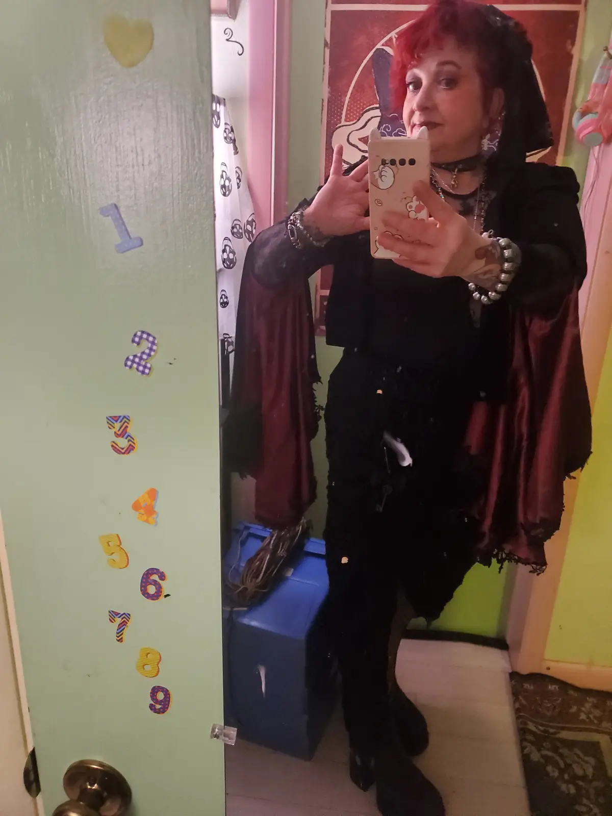 A person with red hair and gothic attire, including a black hooded outfit and cape, takes a mirror selfie while standing…