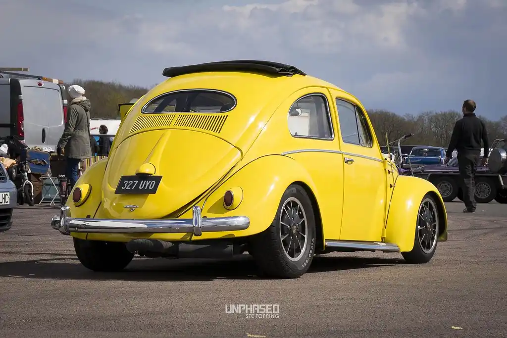 A bright yellow classic Volkswagen Beetle is shown from a rear three-quarter angle at an outdoor car show, with people and…