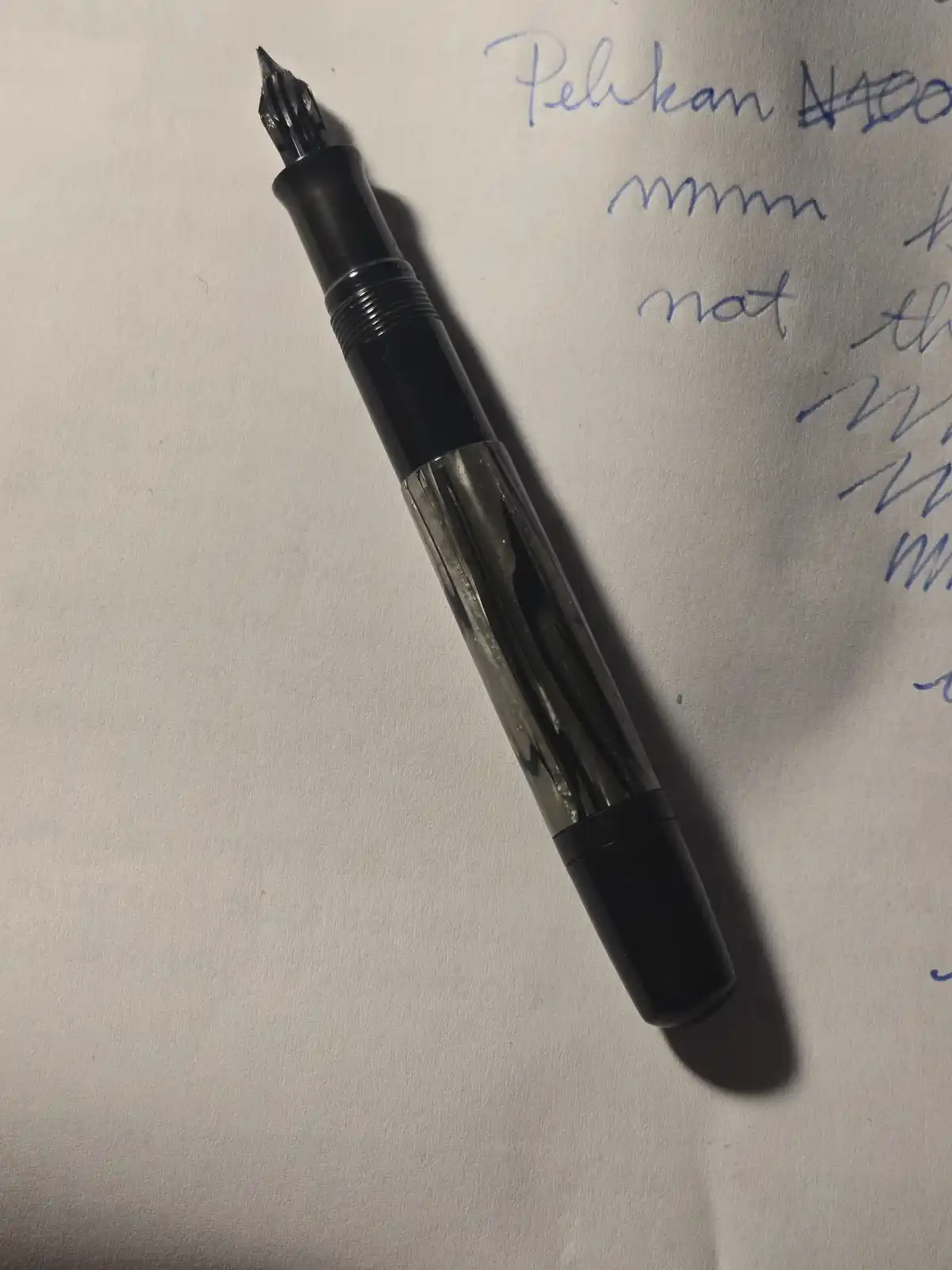 A black fountain pen with a transparent barrel rests on white paper showing handwritten text that reads 'Pelikan N100'…