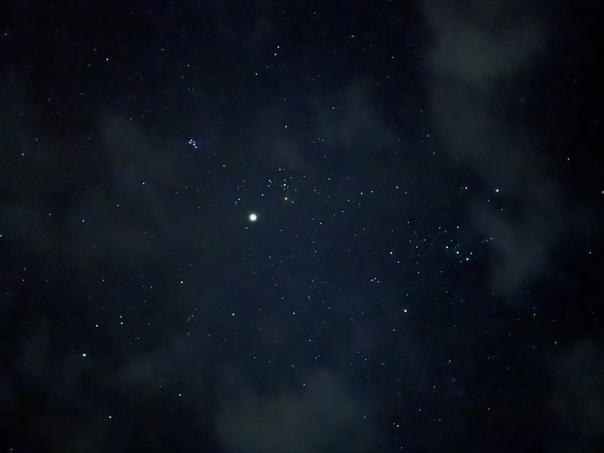 A night sky photo showing numerous stars and faint clouds, with a bright white star prominently visible near the center…