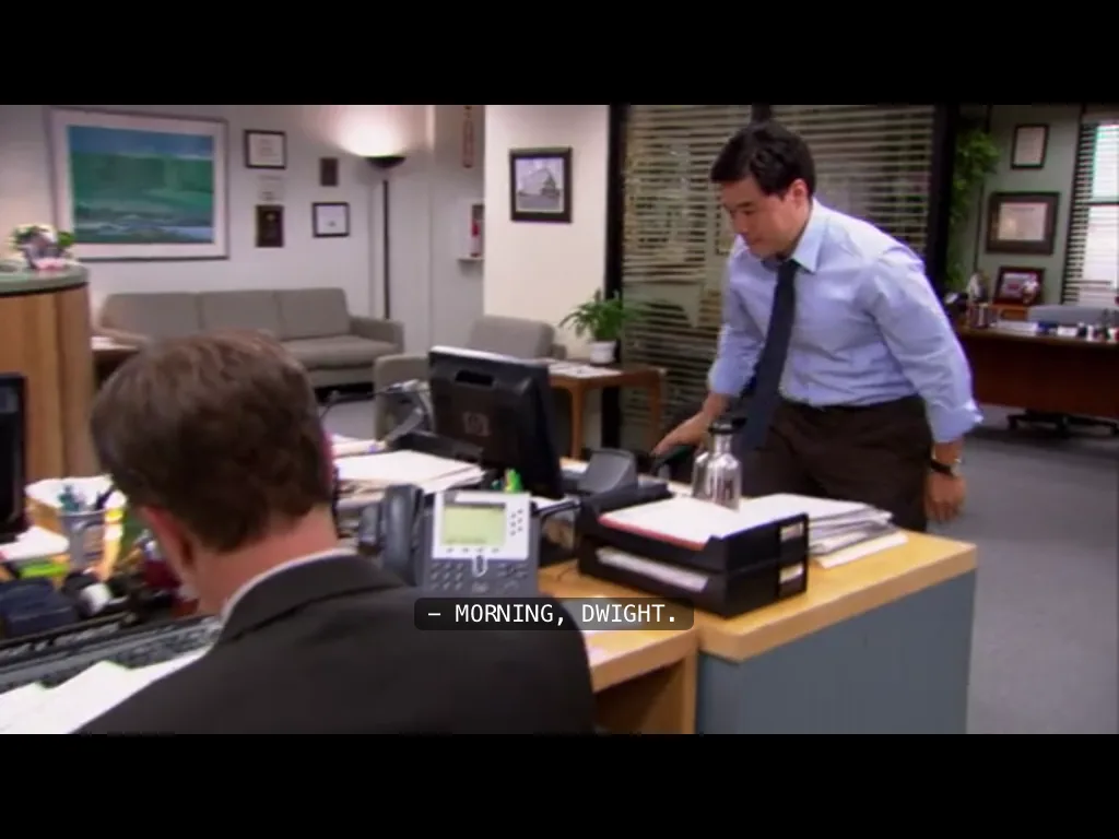A man in a blue shirt and tie leans over a cluttered office desk toward another man seated at the desk, who is seen from…