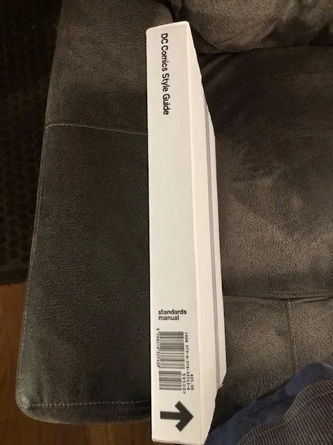 A white book titled 'DC Comics Style Guide' with 'standards manual' and a barcode is shown standing upright on a dark gray…