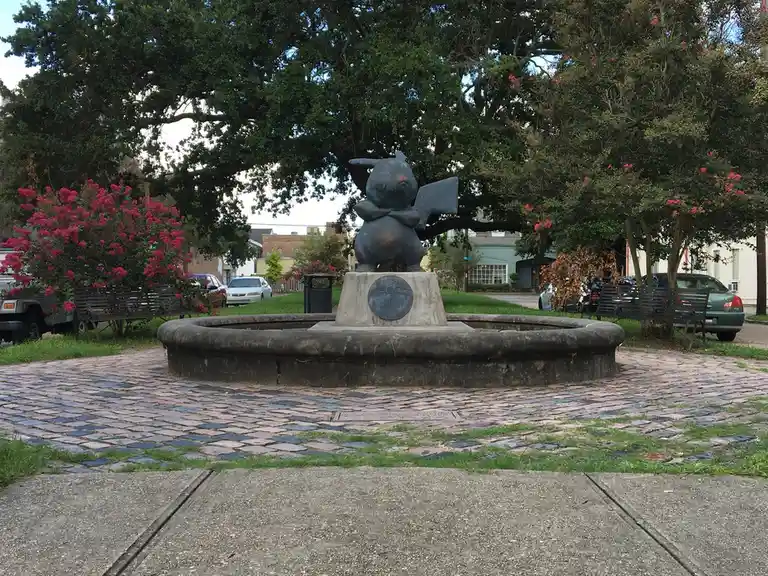 A bronze statue of Pikachu, the iconic Pokémon, is seated atop a stone fountain pedestal in a public park. The statue is…