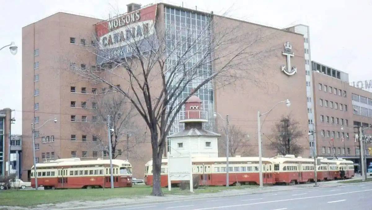 A vintage photo shows a large brick building with a massive 'Molson's Canadian Beer' sign and a giant anchor logo. In front…