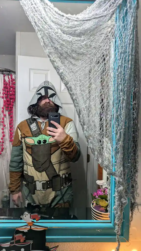 A man with a beard wearing a 'The Mandalorian' onesie with Grogu (Baby Yoda) printed on the front takes a mirror selfie. A…