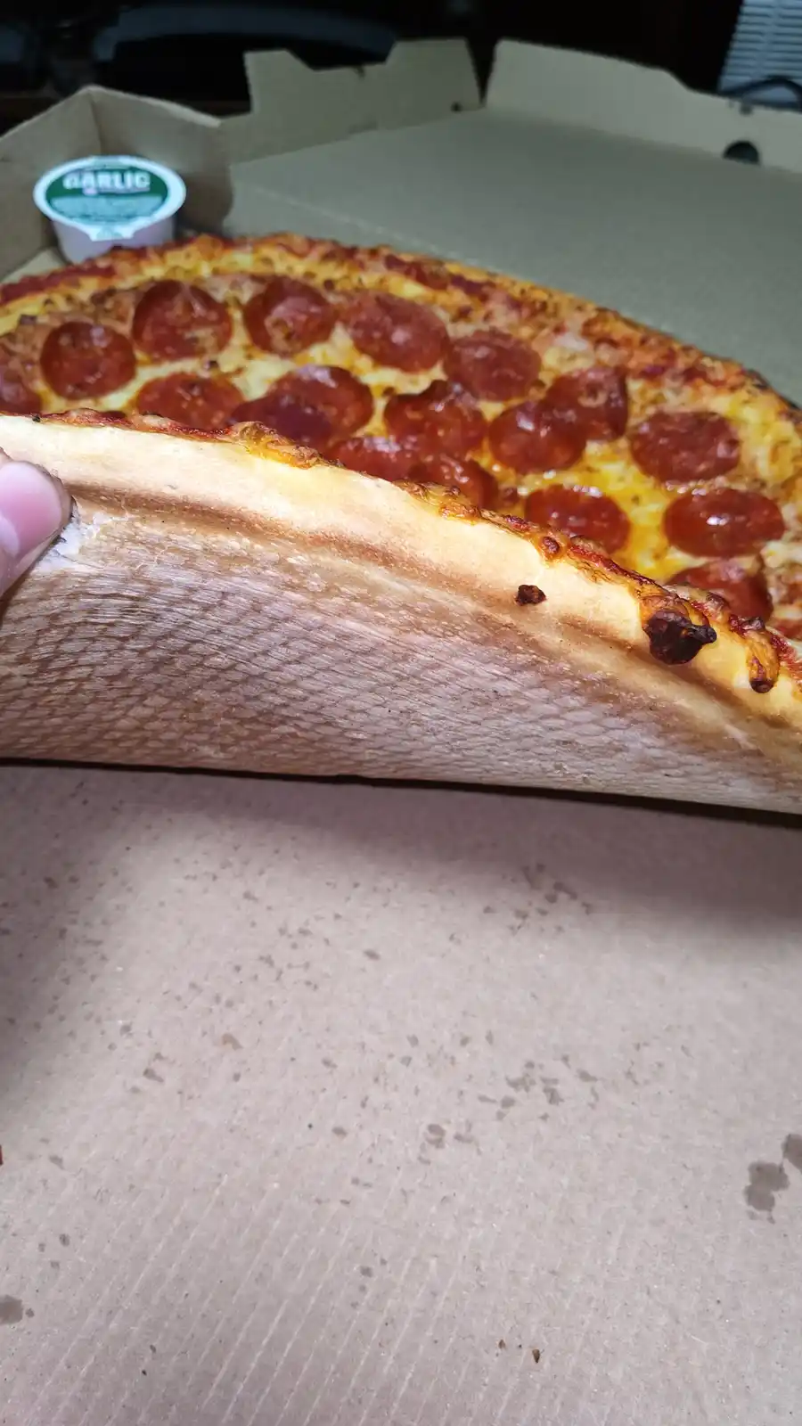 A close-up photo shows a hand lifting a thick slice of pepperoni pizza from a cardboard box, revealing its dense, airy, and…