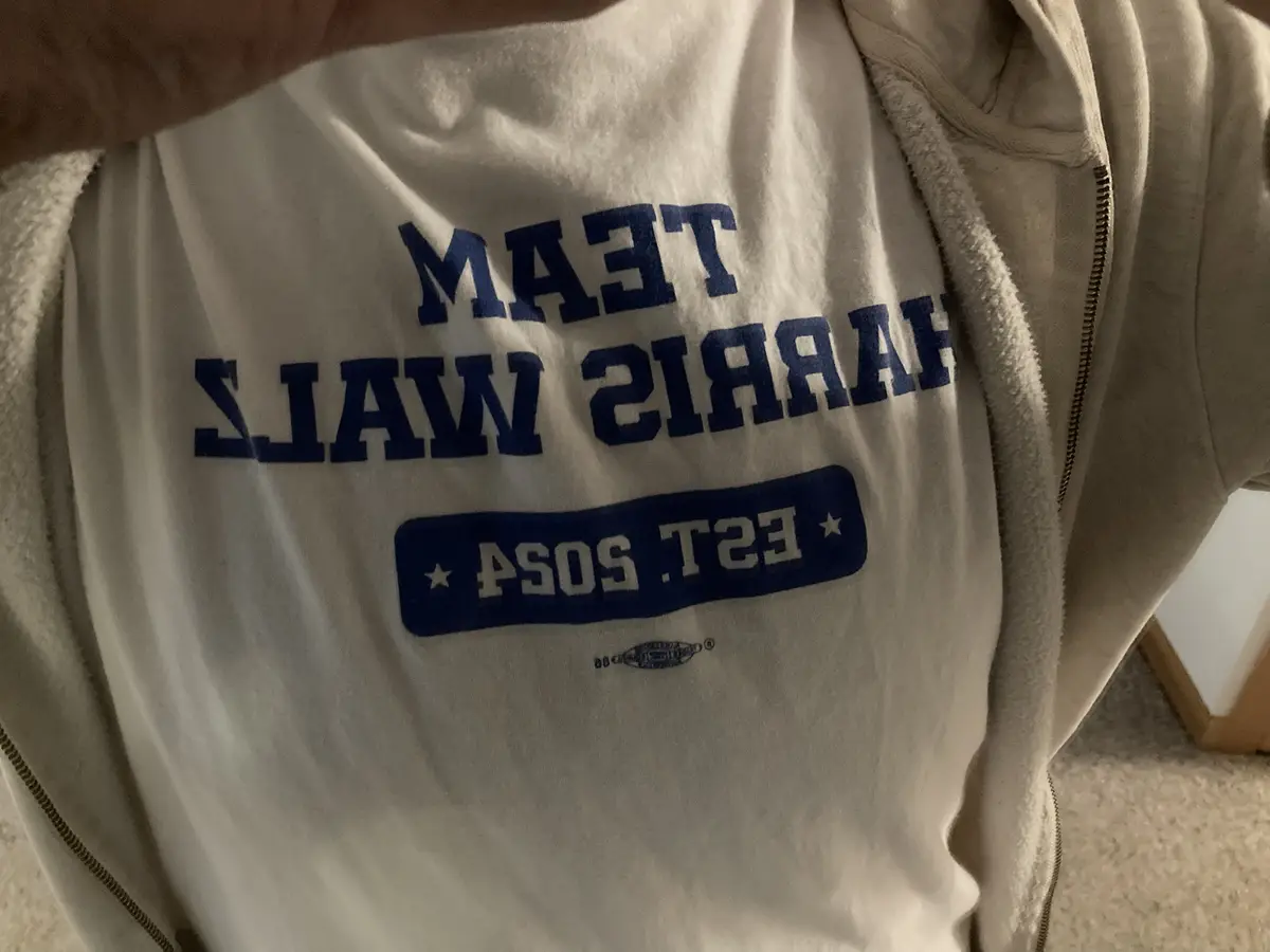 A close-up photo shows a person wearing a white t-shirt under an open beige zip-up hoodie. The t-shirt has bold blue text…