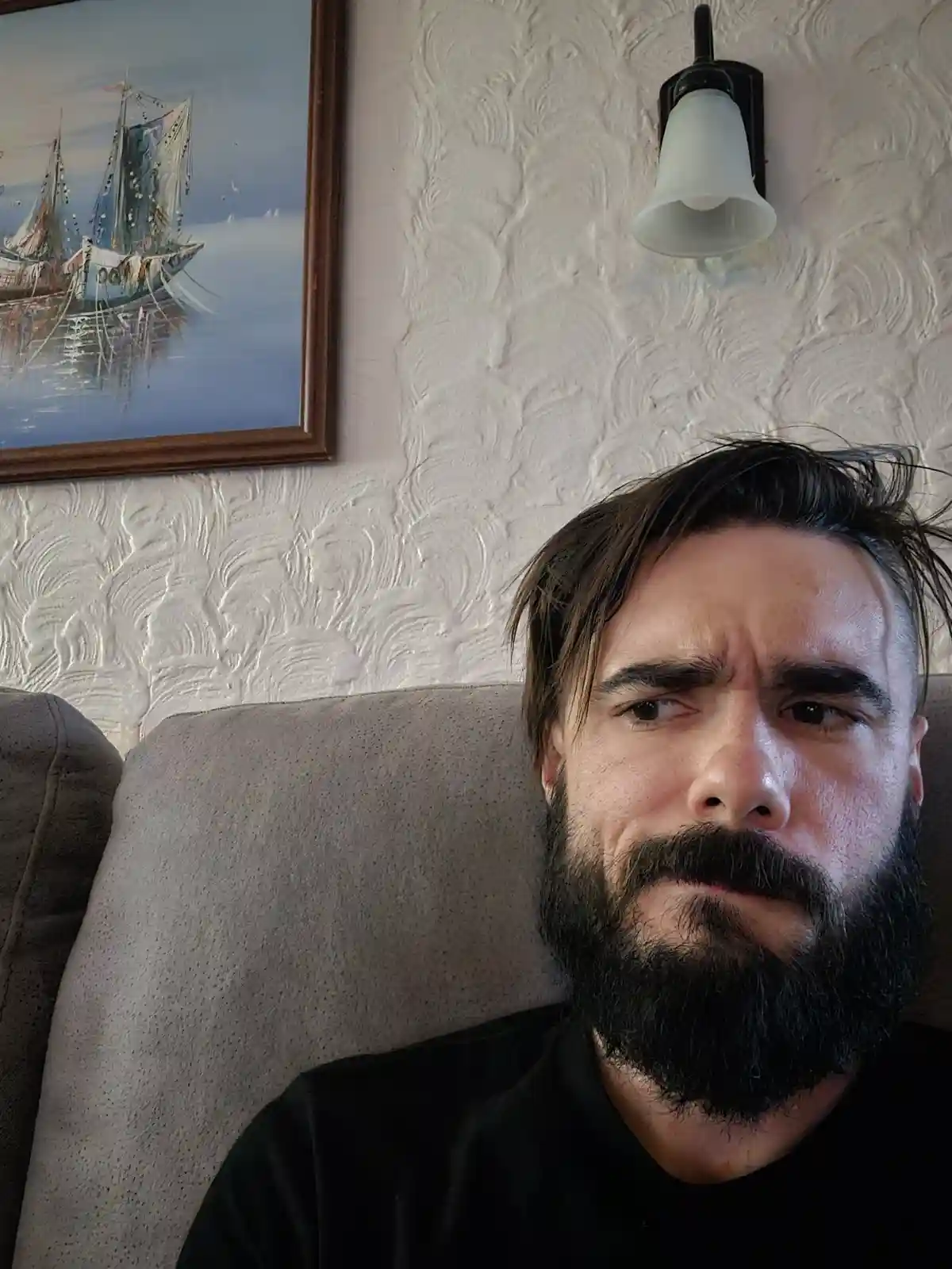 A man with a full beard and dark hair sits on a gray couch, looking directly at the camera with a skeptical, slightly…