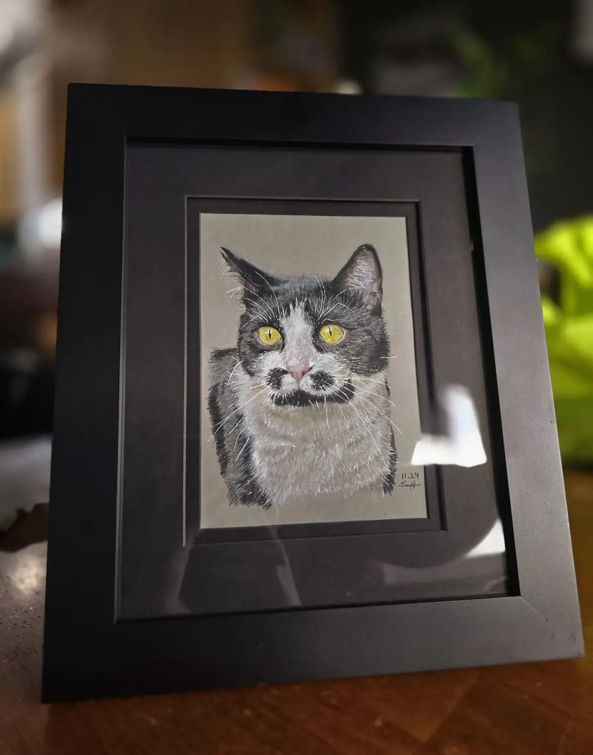A framed portrait drawing of a black and white cat with striking yellow eyes and a distinct black mustache marking on its…