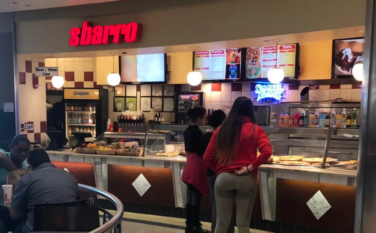A photo of the inside of an Sbarro pizza restaurant, showing the counter with menu boards, a Snapple cooler, and customers…