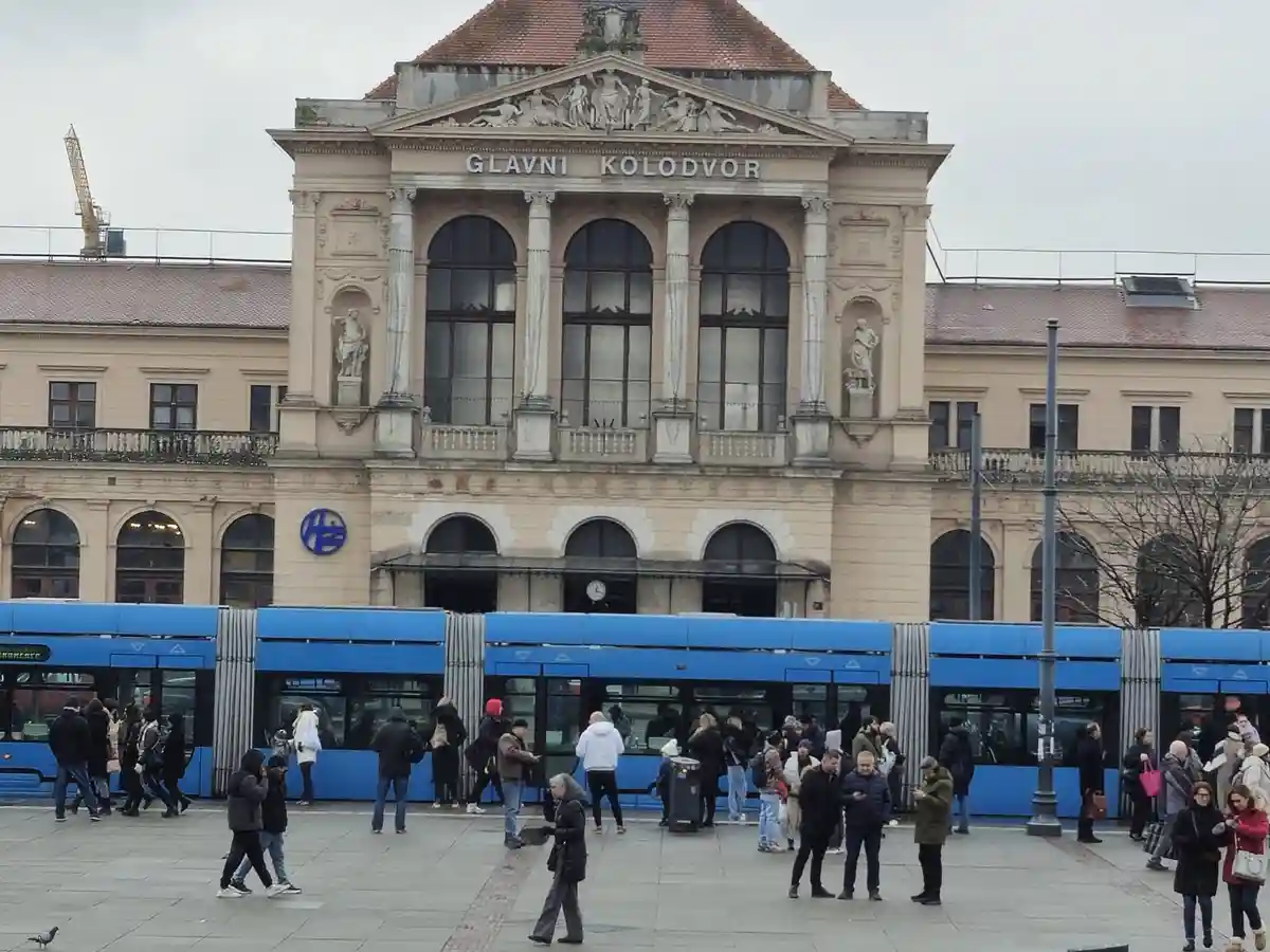 A large, ornate building with the words 'GLAVNI KOLODVOR' above its entrance, with a blue tram stopped in front and many…