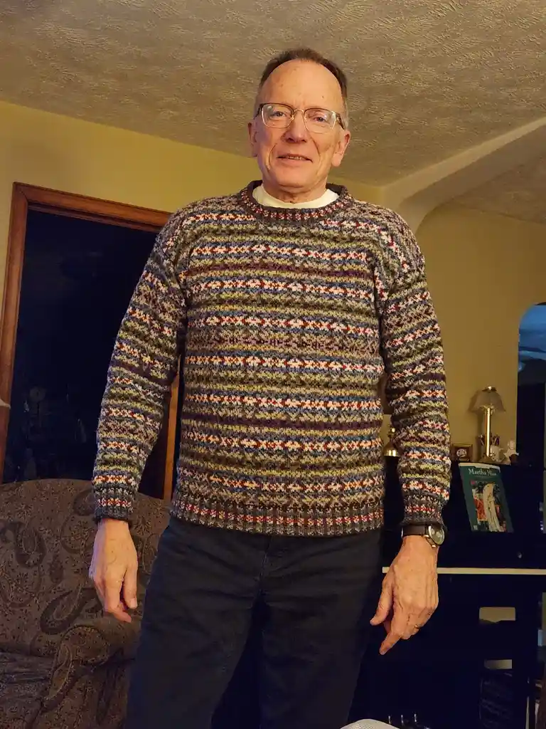 An older man with glasses stands in a warmly lit room wearing a colorful, patterned hand-knitted sweater with horizontal…