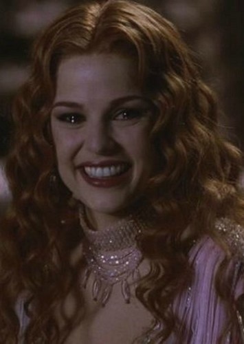 a close-up of a woman with long, curly red hair and a wide, toothy grin, wearing an ornate jeweled choker and a…