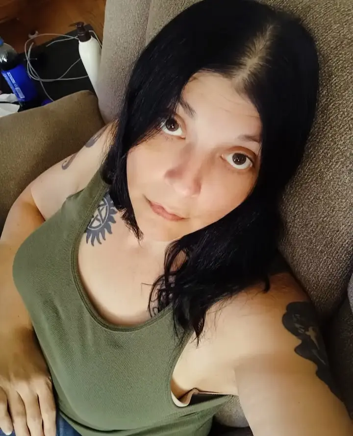 A woman with dark hair and visible tattoos, including a sigil on her neck and a floral design on her right arm, lies on a…