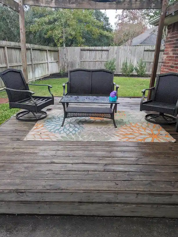 An outdoor wooden deck features a black wicker patio set including two swivel chairs and a loveseat with a glass-top coffee…