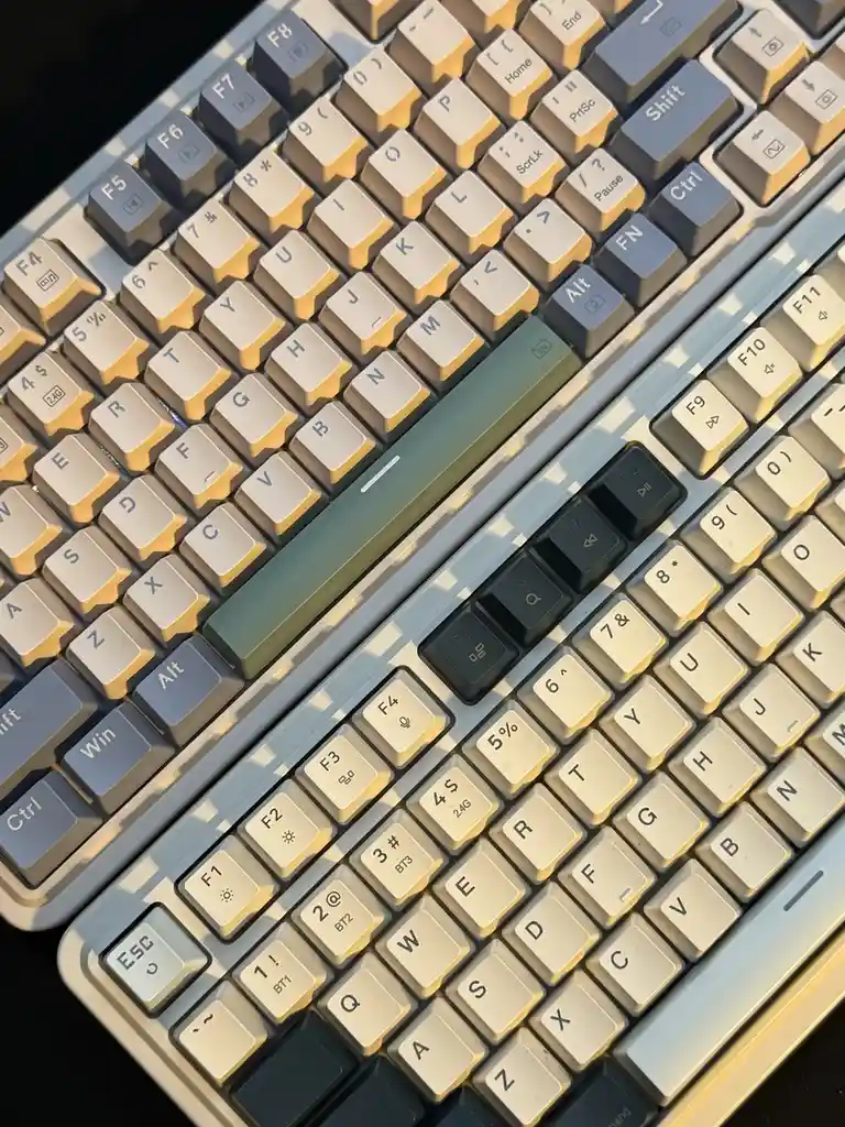 Two mechanical keyboards are placed side by side, angled diagonally, with sunlight casting shadows across their keys. The…