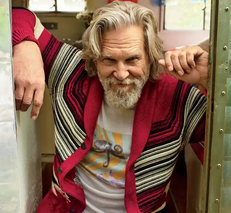 A man with long blonde hair and a beard, wearing a red cardigan over a t-shirt with an octopus graphic, leans against a…