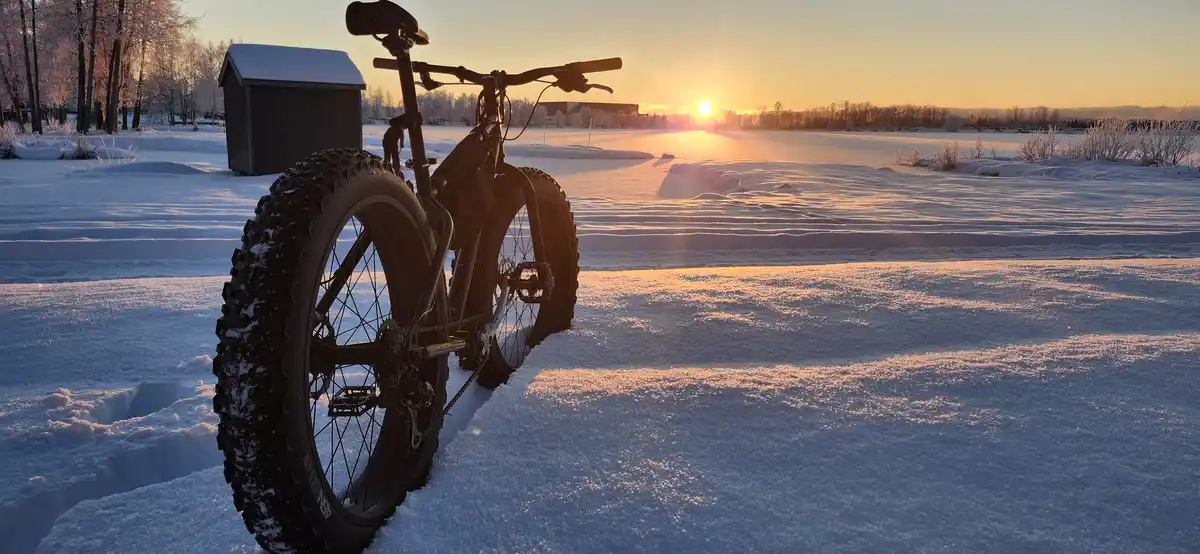 A fatbike with thick, treaded tires stands upright in deep snow, silhouetted against a golden sunset over a frozen…