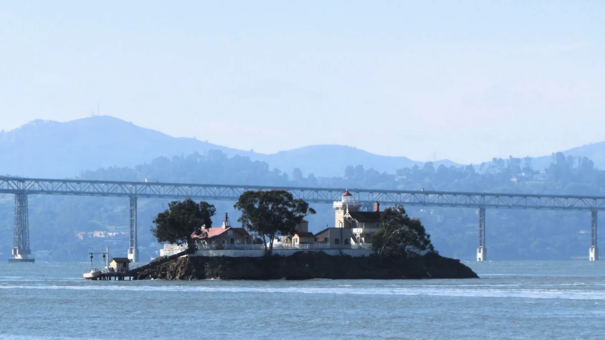 A scenic view of a small island with a lighthouse and several buildings, situated in a bay with a large suspension bridge…
