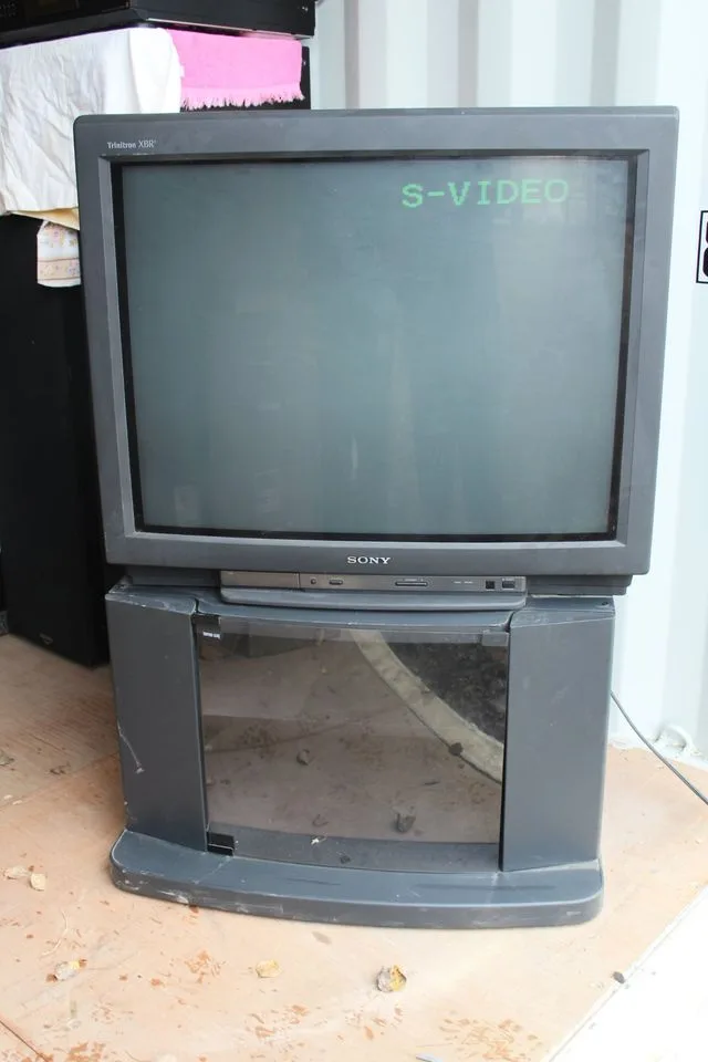 An old, dusty Sony Trinitron XBR television sits on a dirty stand in a cluttered room, its screen displaying the text…
