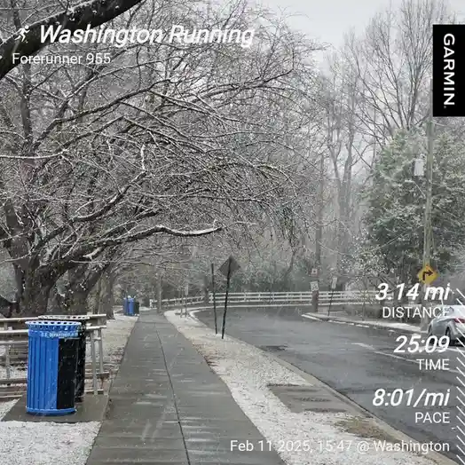 A snowy sidewalk scene with bare trees covered in snow, a blue trash can, and a road with a car and a white fence in the…