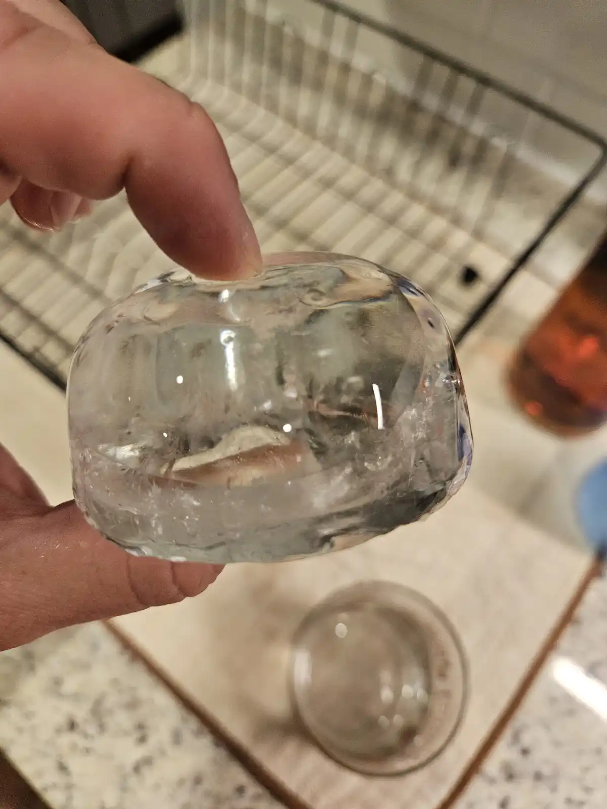 A hand holds a large, clear, homemade ice cube that contains a small, visible worm trapped inside. The background shows a…