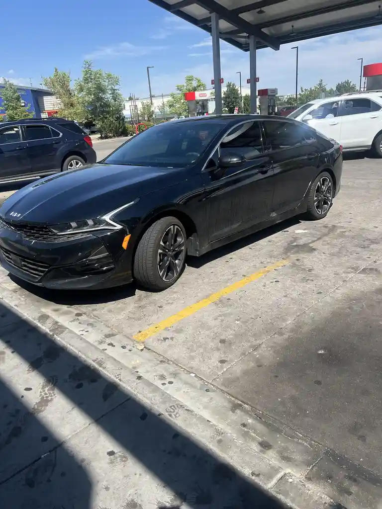 A black Kia sedan is parked at a gas station under a canopy, with visible fuel pumps in the background. The car is…