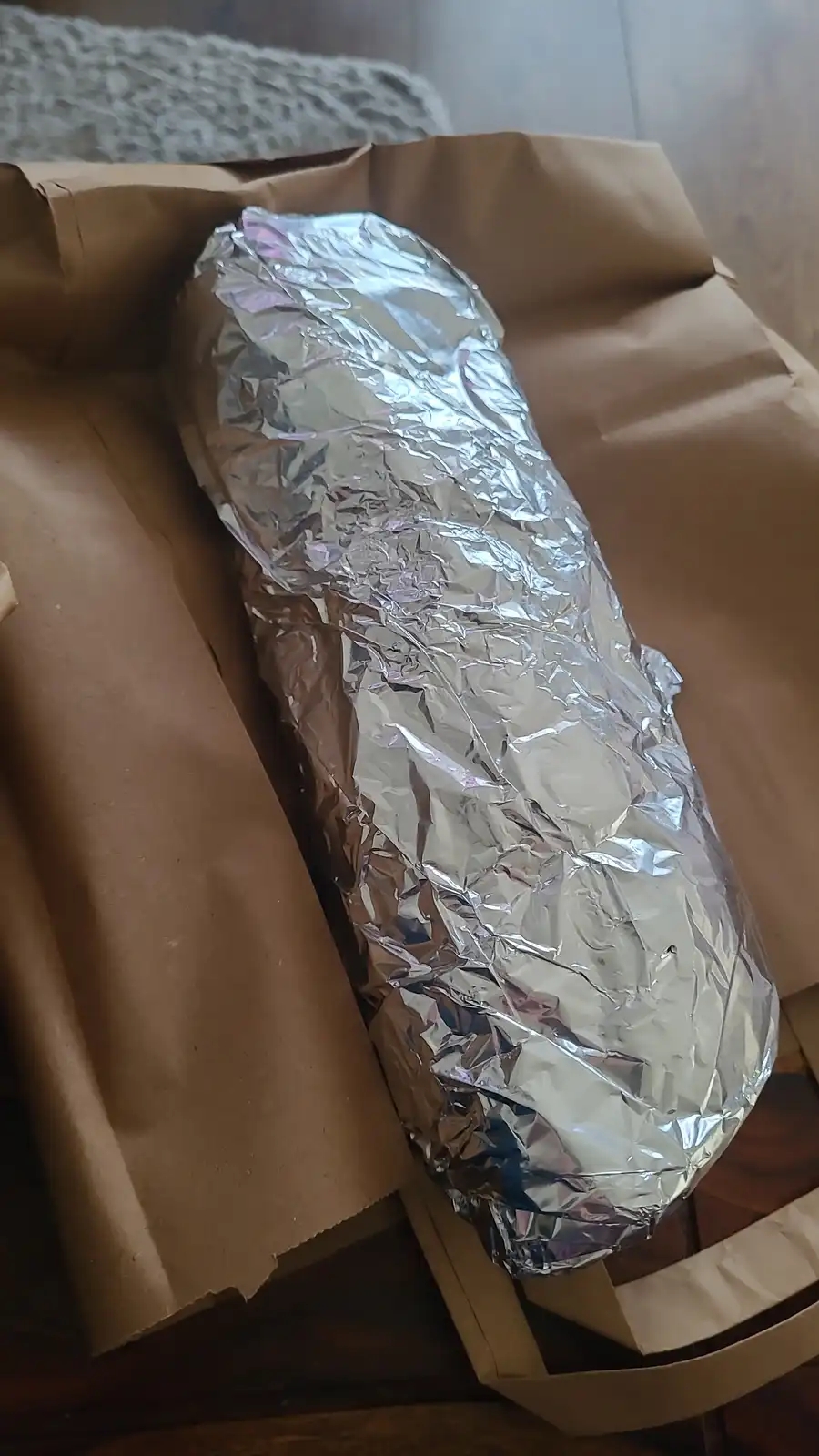 A long, cylindrical food item, likely a burrito or wrap, is tightly wrapped in crinkled aluminum foil and rests inside an…