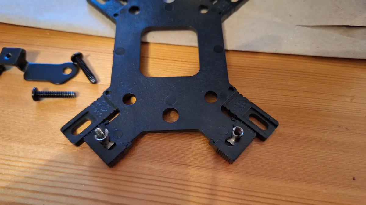 A close-up photo shows a black plastic mounting bracket with several holes and slots, lying on a wooden surface. Two screws…