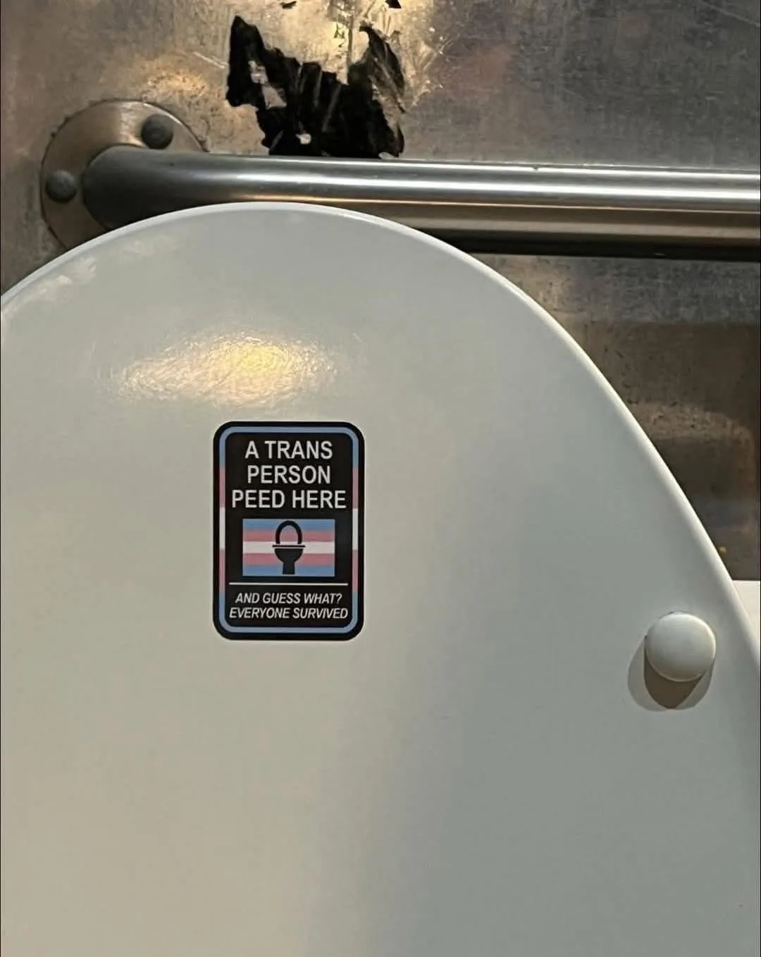 A close-up photo shows a sticker on the lid of a public toilet stall. The sticker has a black border, white text, and a…