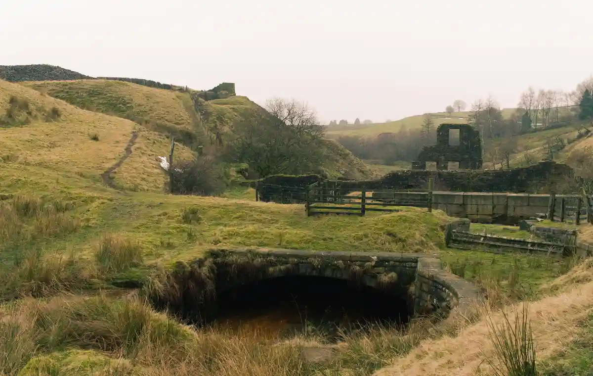 A rural landscape featuring a stone bridge over a small stream, with grassy hills and ancient stone ruins in the background…