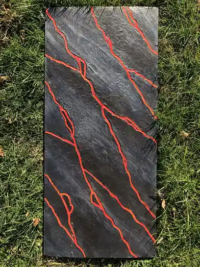 A rectangular black stone slab with bright orange-red painted cracks lies on green grass, mimicking a volcanic rock. The…
