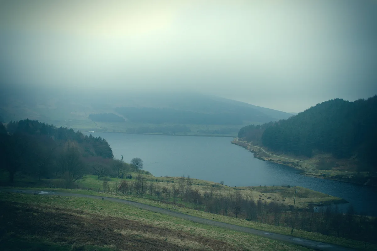 A misty, atmospheric landscape photo of a large lake surrounded by rolling hills and forests, with a winding road in the…