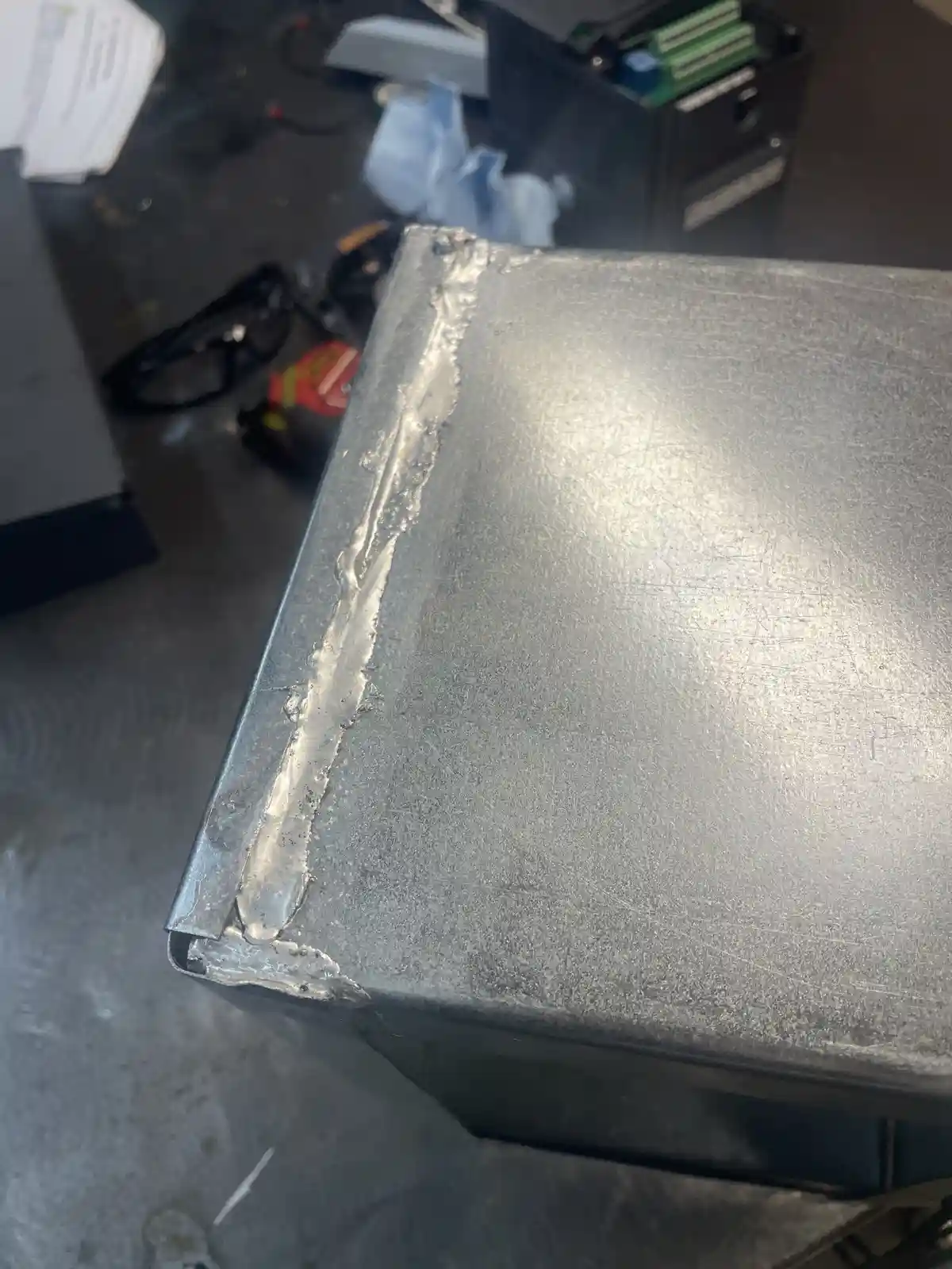 A close-up photo shows a rough, uneven weld seam along the edge of a metal box, with a cluttered workshop background…