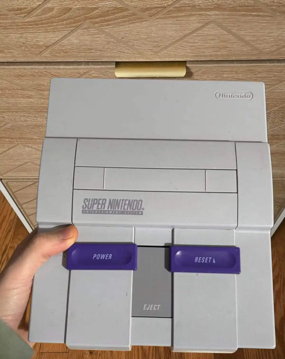 A hand holds a grey Super Nintendo Entertainment System console, with its purple 'POWER' and 'RESET' buttons clearly…
