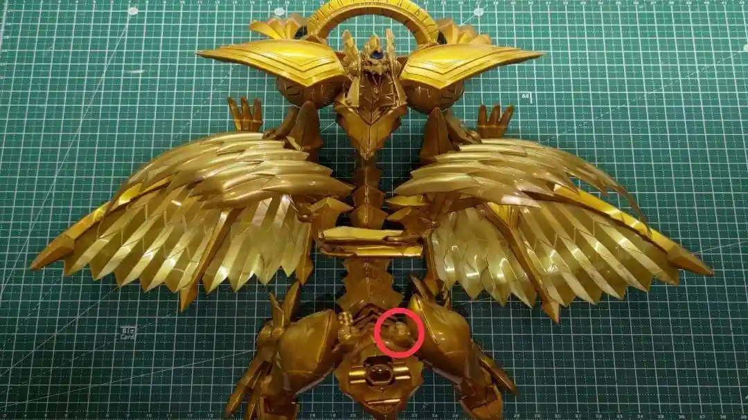 A golden, intricately detailed robot model with large, feathered wings is displayed on a green cutting mat. A red circle…