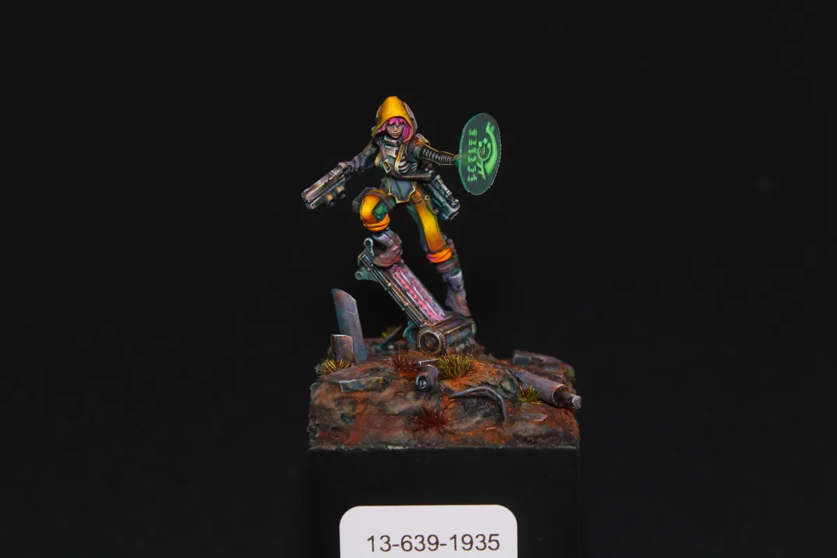 A painted miniature figure of a futuristic warrior with pink hair, a yellow hood, and a green glowing shield stands on a…
