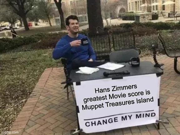 A man sits at a small outdoor table with a sign that reads 'Hans Zimmers greatest Movie score is Muppet Treasures Island'…