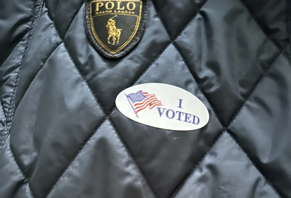 A close-up of a black quilted jacket with a gold and black Polo Ralph Lauren logo patch and an oval 'I VOTED' sticker with…
