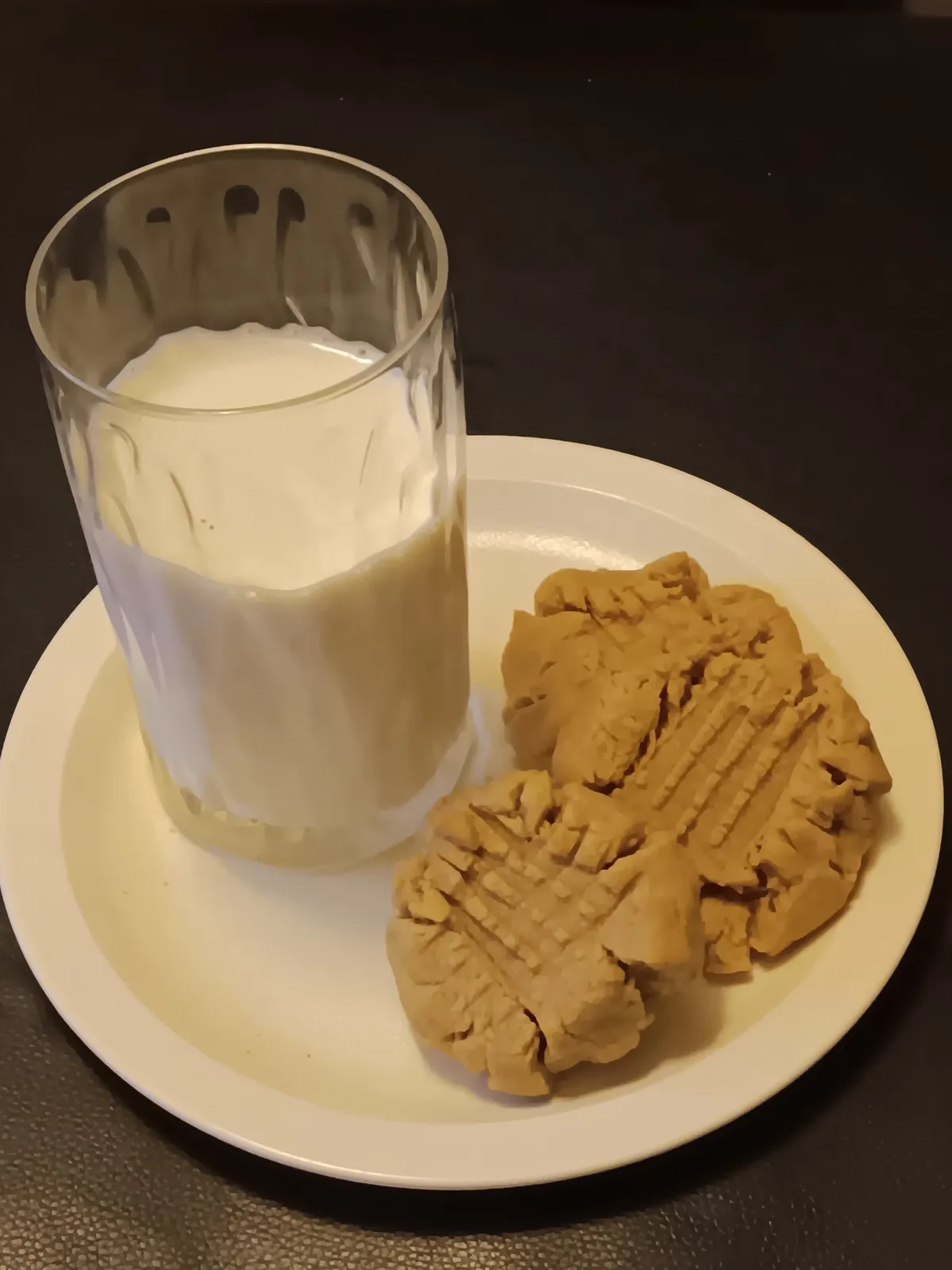 A white plate holds two peanut butter cookies with crisscross patterns and a glass of milk, both resting on a dark surface…