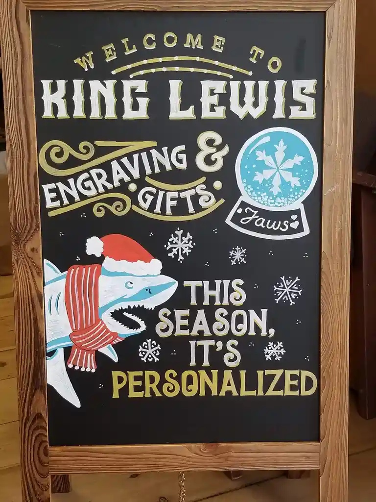 A hand-painted chalkboard sign for 'King Lewis Engraving & Gifts' features festive holiday elements including a shark…