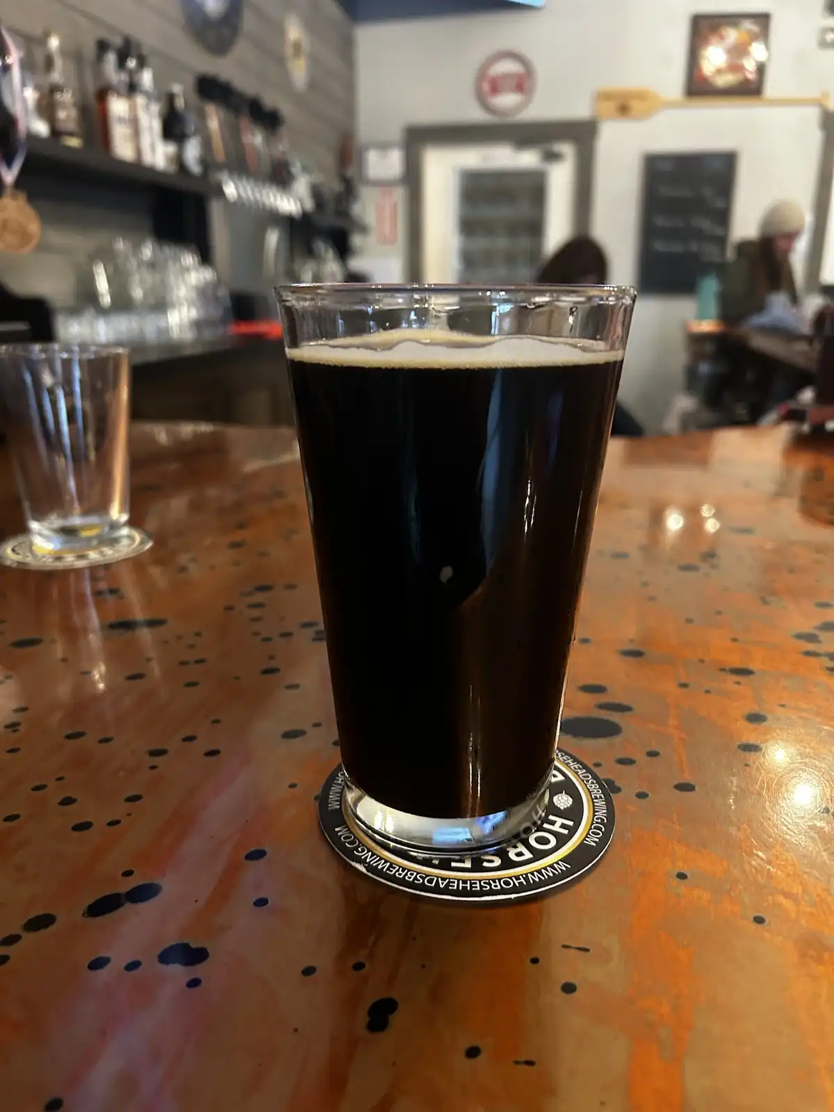 A tall glass of dark beer with a light head sits on a speckled wooden bar top, resting on a circular coaster that reads…