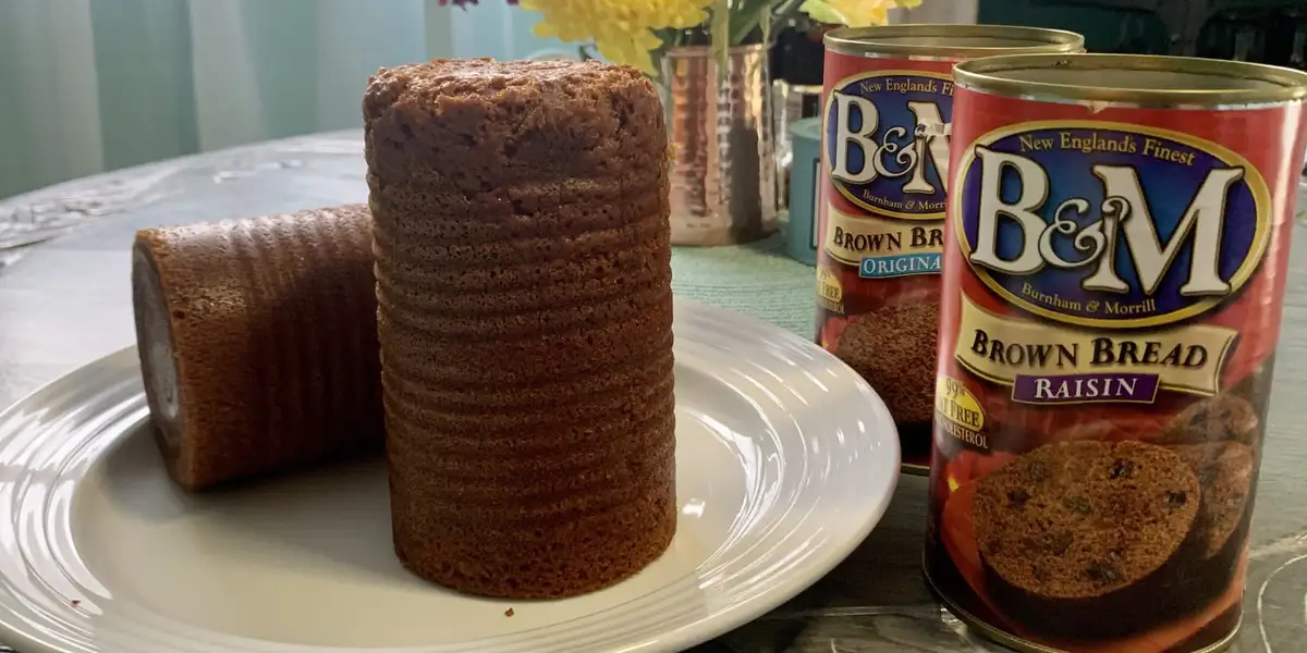 A tall, cylindrical stack of brown bread slices sits on a white plate next to a can of B&M Brown Bread Raisin, with another…