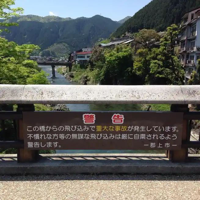A brown warning sign in Japanese is mounted on a concrete bridge railing overlooking a river with mountains and buildings in…