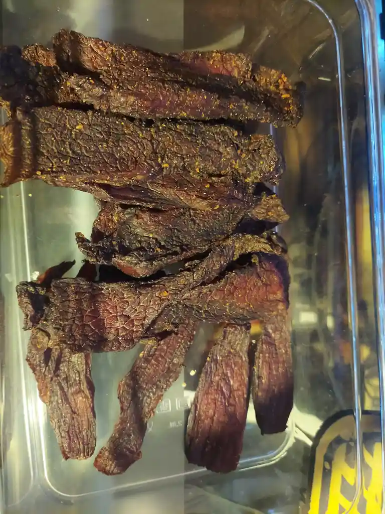A clear plastic container holds a pile of dark brown, textured beef jerky strips seasoned with orange spices. The text…