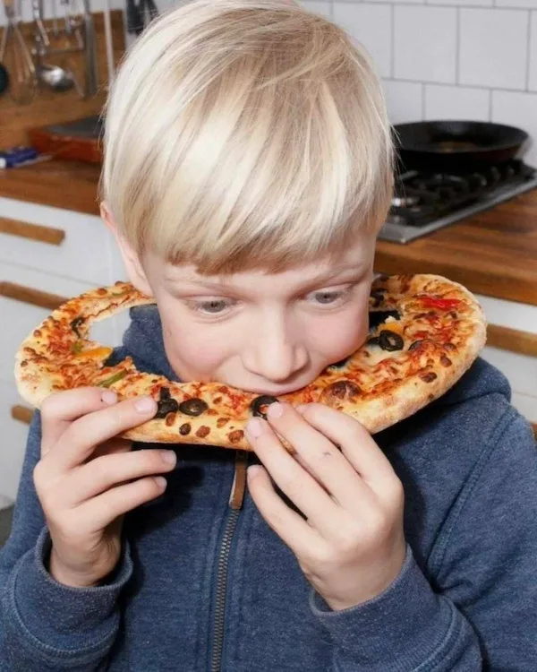 A young blond boy in a blue zip-up hoodie holds a small, round pizza with both hands, positioning it around his neck like a…