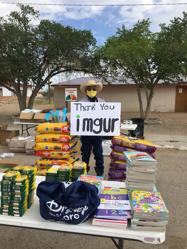 A man wearing a cowboy hat, sunglasses, and a yellow face mask stands outdoors holding a white sign that reads 'Thank You…