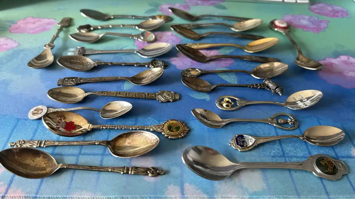 A collection of various souvenir spoons with different designs and engravings, laid out on a blue and green floral…